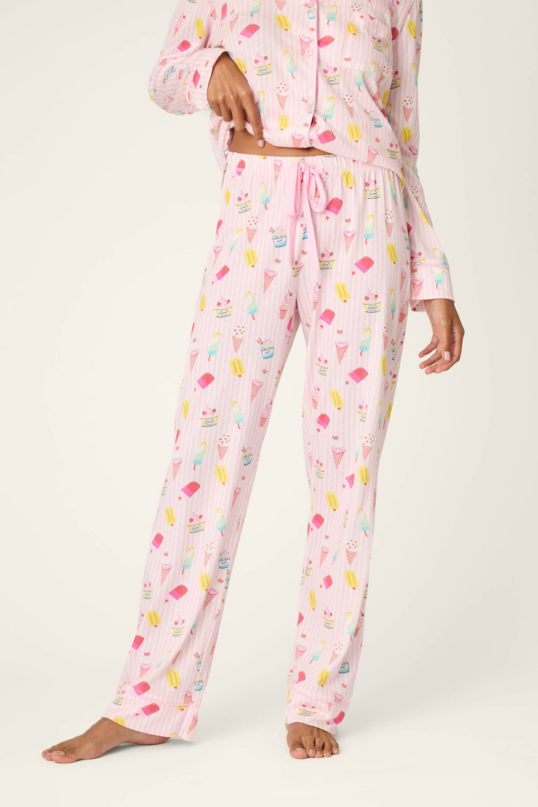 A person stands barefoot wearing P.J. Salvage Playful Prints Sugar Rush Pajama Pants, featuring a fun dessert print. The image showcases the lower half of their body in these pastel pink pajama pants.