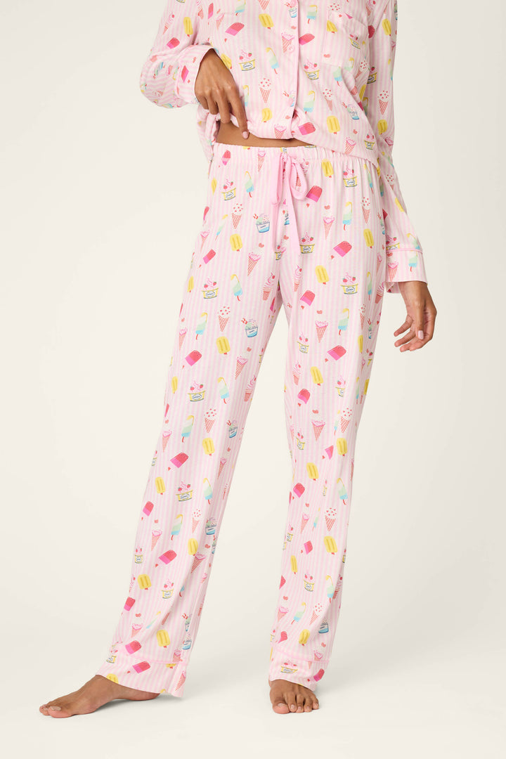 A person stands barefoot wearing P.J. Salvage Playful Prints Sugar Rush Pajama Pants, featuring a fun dessert print. The image showcases the lower half of their body in these pastel pink pajama pants.