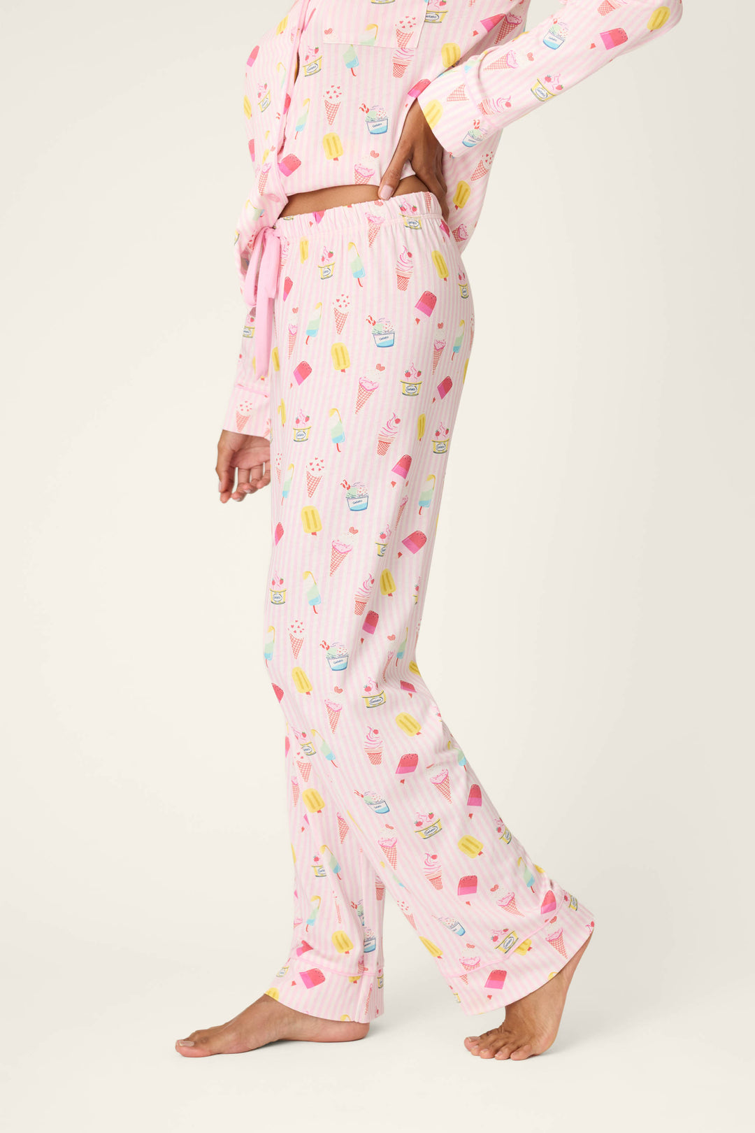 A person stands barefoot in P.J. Salvage Playful Prints Sugar Rush Pajama Pants and a matching cotton-modal long-sleeve top, both in light pink, against a plain, light background.