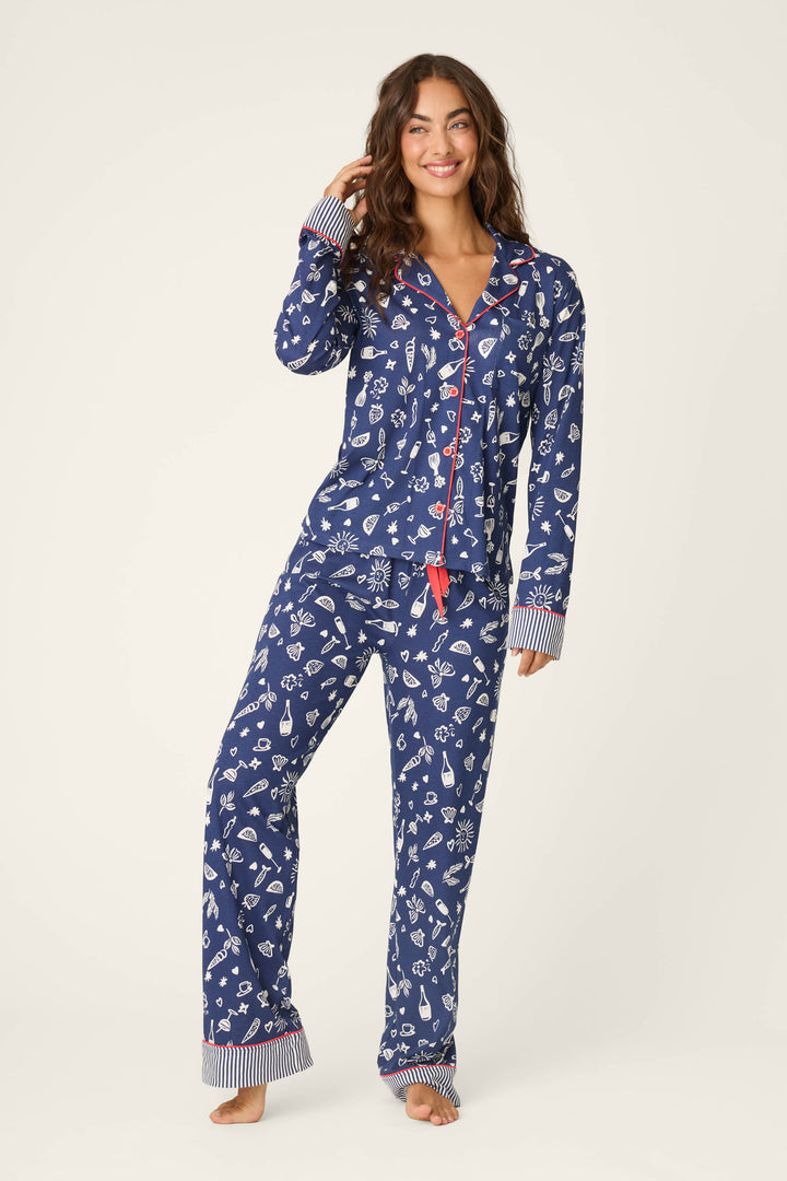 A woman stands barefoot, smiling, wearing the P.J. Salvage Playful Prints Al Fresco Bistro Long PJ Set—an Italian-inspired navy blue pajama set with white winter patterns and red accents—crafted from soft cotton-modal jersey, against a plain light background.