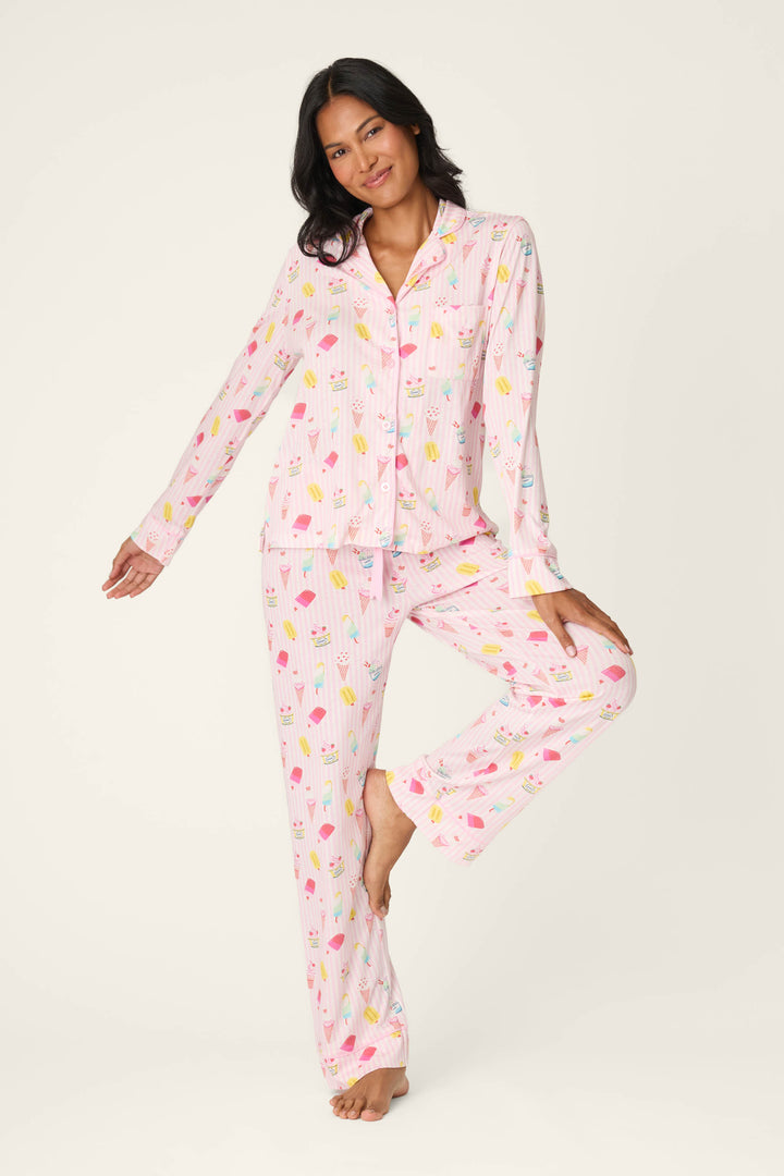 A woman models the P.J. Salvage Playful Prints Sugar Rush Long PJ Set—a pink, long-sleeved pajama set featuring a colorful ice cream print—as she stands barefoot, smiling with one arm out against a plain light background.