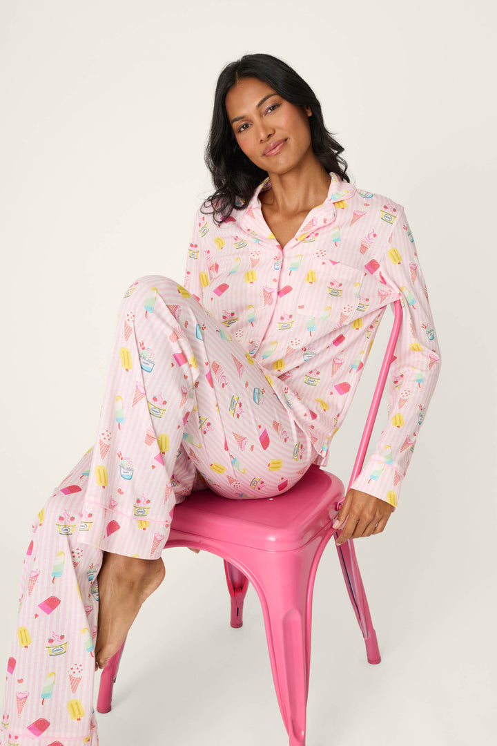 A woman with long dark hair sits sideways on a pink chair, smiling softly at the camera as she models the P.J. Salvage Playful Prints Sugar Rush Long PJ Set—cotton-modal jersey pajamas featuring colorful ice cream and popsicle designs—against a white background.