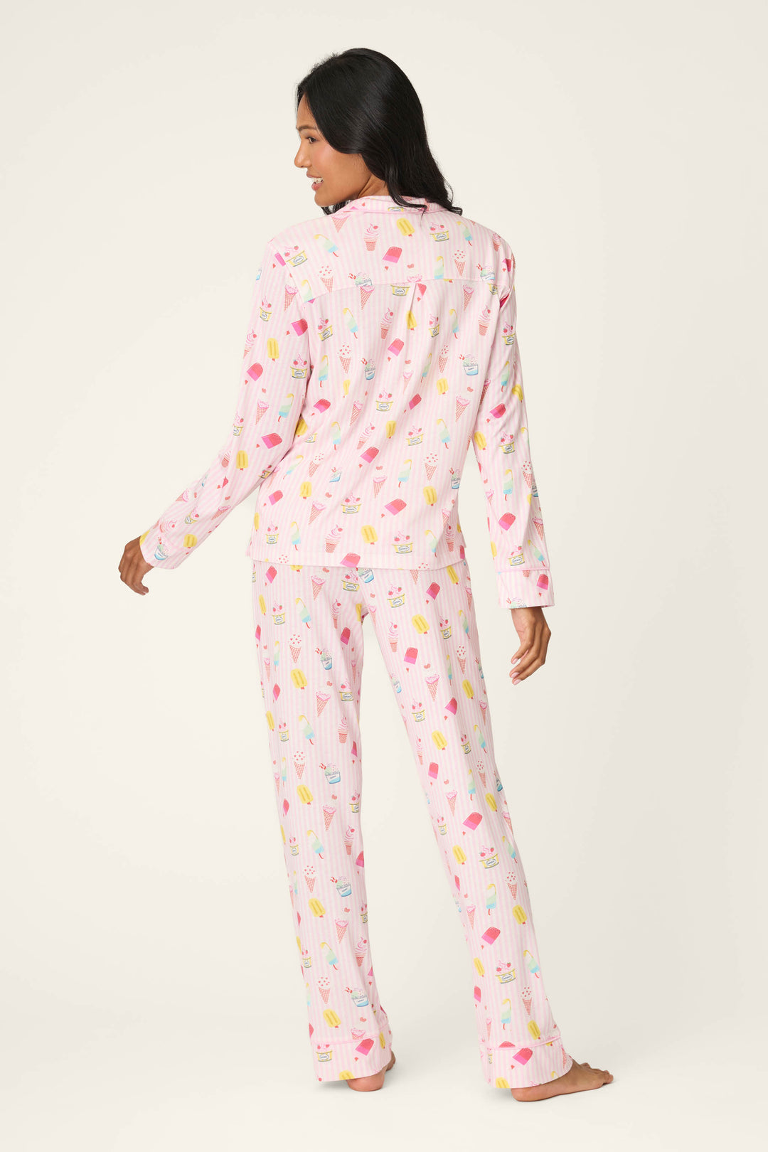 A woman stands barefoot, facing away, wearing the P.J. Salvage Playful Prints Sugar Rush Long PJ Set in light pink with colorful ice cream cones, made from soft cotton-modal jersey, against a plain off-white background.