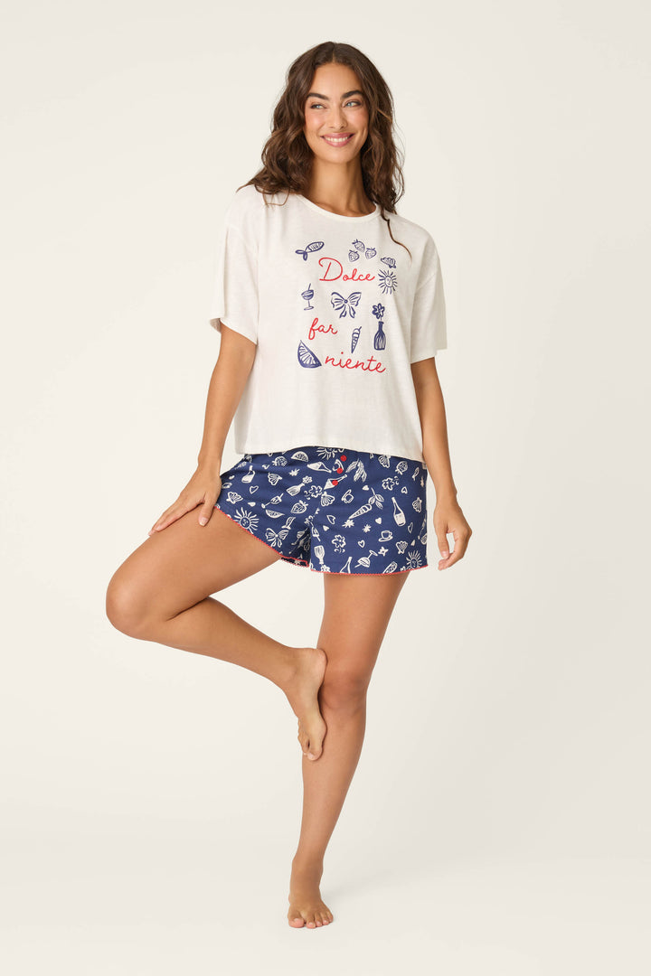 A woman with long brown hair stands on one leg, smiling, wearing the P.J. Salvage Playful Prints Dolce Far Niente Short PJ Set—Italian-inspired, with a colorful design and blue patterned shorts—against a plain light background.