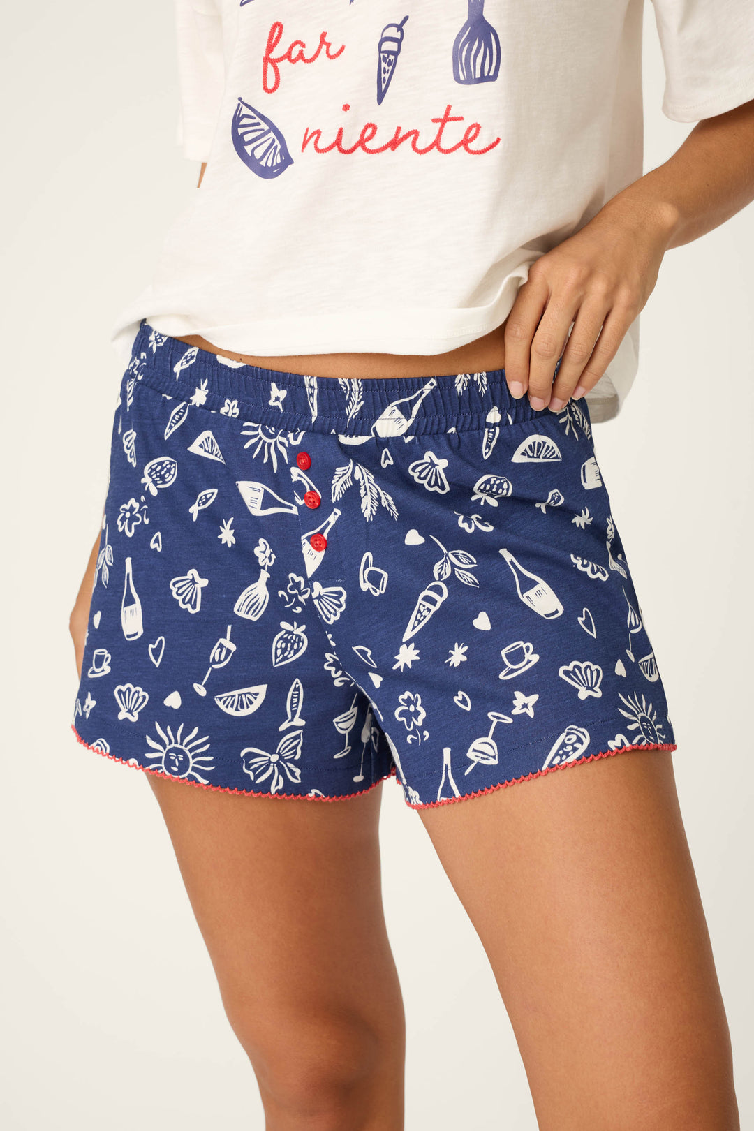 A person wears the P.J. Salvage Playful Prints Dolce Far Niente Short PJ Set, featuring a white tee with red text and blue shorts with white summer-themed prints and red trim—ideal for relaxing in style.