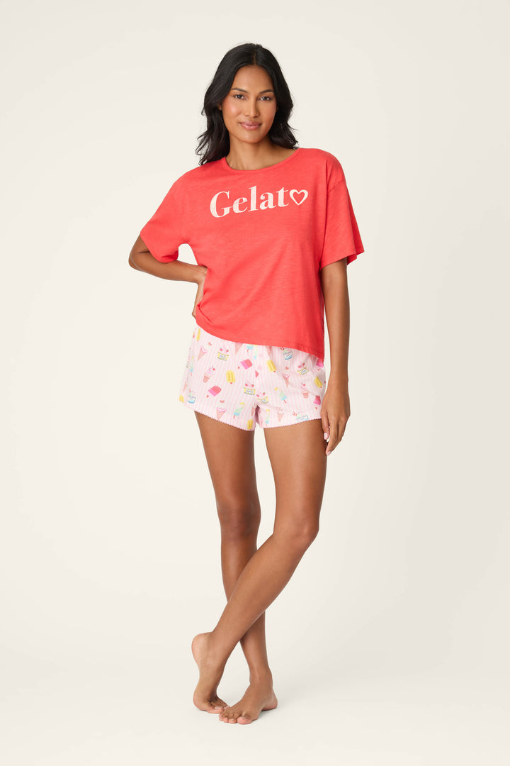 A woman stands barefoot in a relaxed pose, wearing the P.J. Salvage Playful Prints Gelato Love Short PJ Set—a red T-shirt and light pink shorts with a vibrant ice cream print—perfect for stylish lounging against a plain background.
