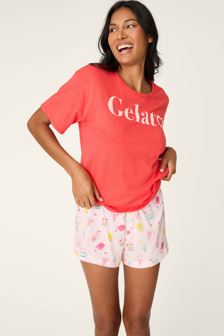 A smiling woman stands with one hand on her hip, wearing the P.J. Salvage Playful Prints Gelato Love Short PJ Set—coral T-shirt and pink ice cream print shorts—an ideal choice for comfy Amalfi Coast loungewear.