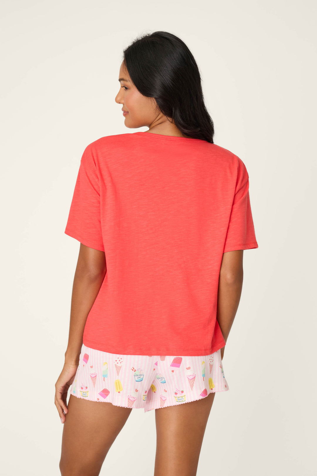 A woman with long dark hair faces away, wearing a relaxed fit Playful Prints Gelato Love Graphic T-Shirt by P.J. Salvage and white shorts with colorful ice cream and popsicle prints, set against a plain light background.