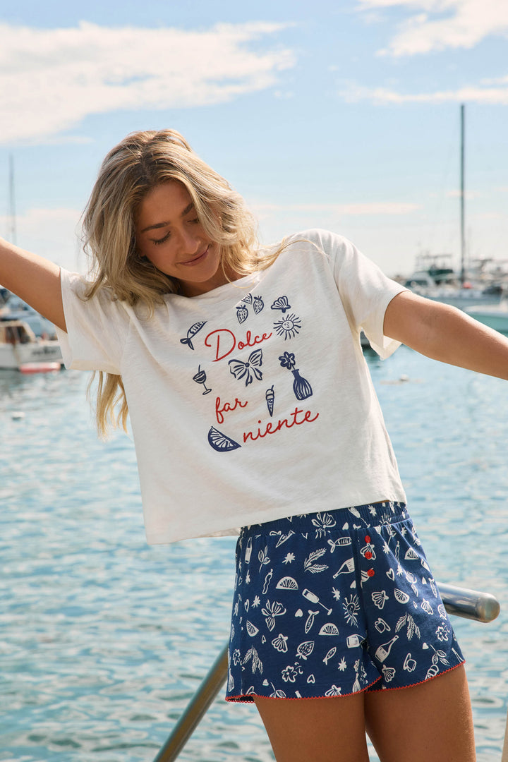 A smiling woman by a marina raises her arms, wearing the P.J. Salvage Playful Prints Dolce Far Niente Graphic T-Shirt and blue sea-print shorts. Boats shimmer behind her, capturing the carefree spirit of la dolce vita under a partly cloudy sky.