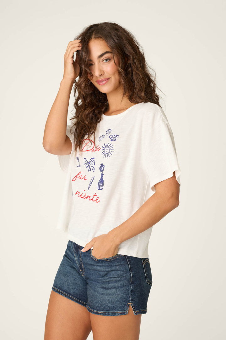 A woman with long curly hair smiles, touching her hair while wearing a P.J. Salvage Playful Prints Dolce Far Niente Graphic T-Shirt and denim shorts, standing against a plain light background.