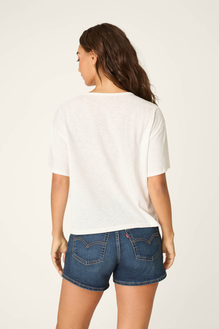 A woman with long brown hair stands with her back to the camera, wearing the P.J. Salvage Playful Prints Dolce Far Niente Graphic T-Shirt and blue denim shorts, evoking the relaxed spirit of la dolce vita against a light backdrop.