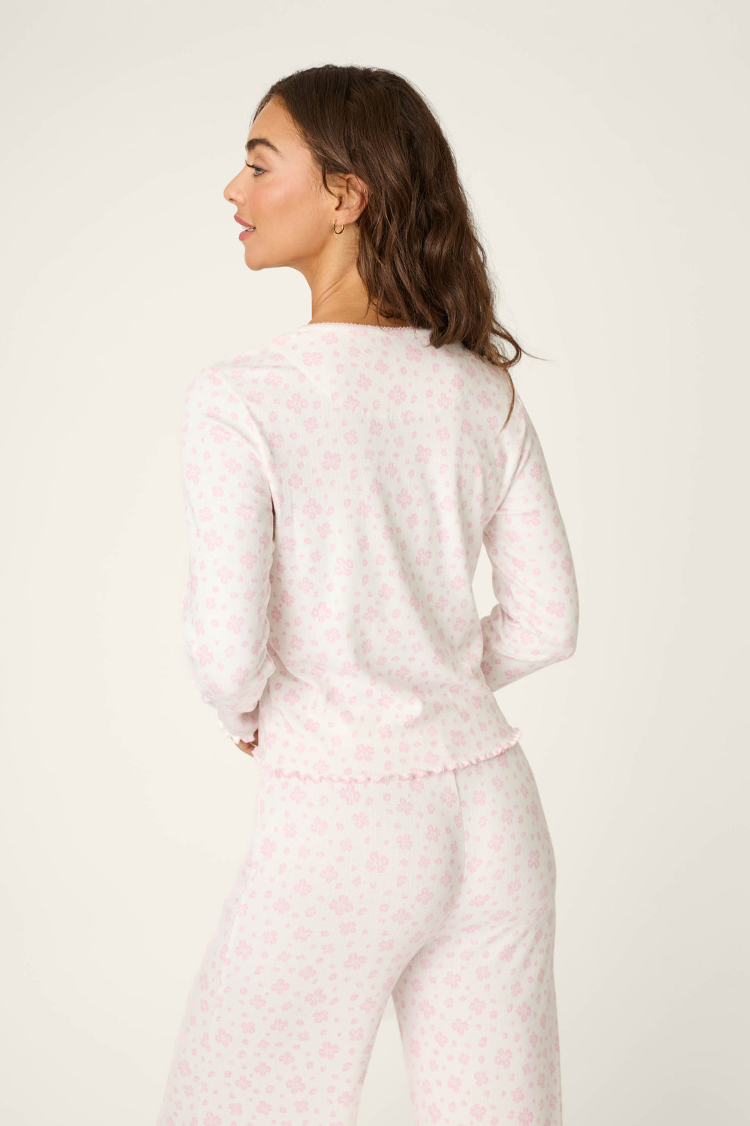A woman with long brown hair faces away, wearing the P.J. Salvage Pretty Petals Sweet Pink Floral Print Long Sleeve Top and matching loose-fitting pants. The pajama set features a delicate floral pattern on a light background.