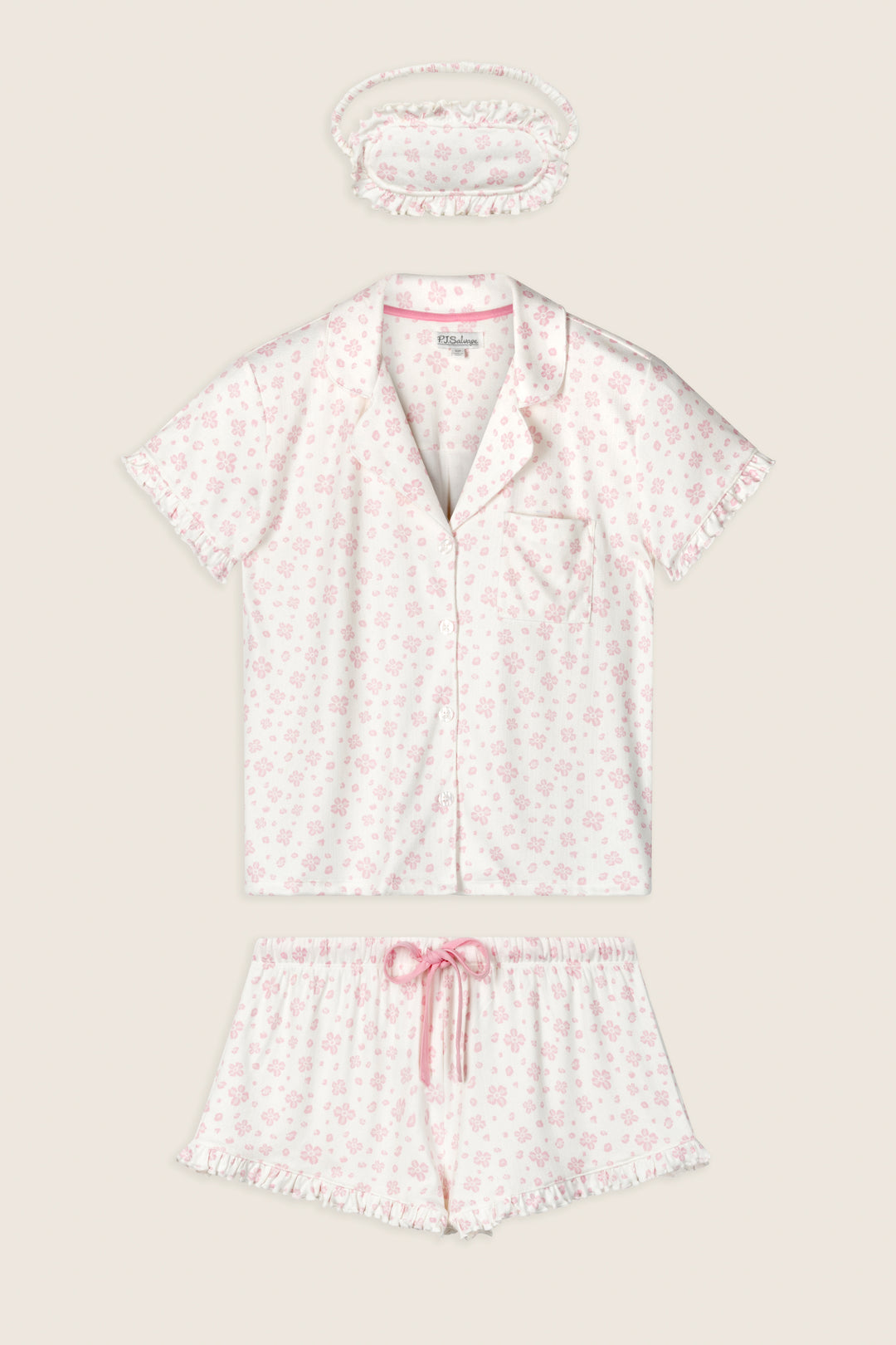 The P.J. Salvage Pretty Petals Sweet Pink Floral Print 3-Piece PJ Set includes a short-sleeve button-up top, drawstring shorts, and a matching scrunchie in white with light pink floral print and ruffle trim.
