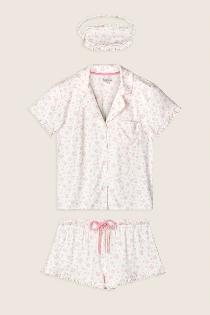The P.J. Salvage Pretty Petals Sweet Pink Floral Print 3-Piece PJ Set includes a short-sleeve button-up top, drawstring shorts, and a matching scrunchie in white with light pink floral print and ruffle trim.