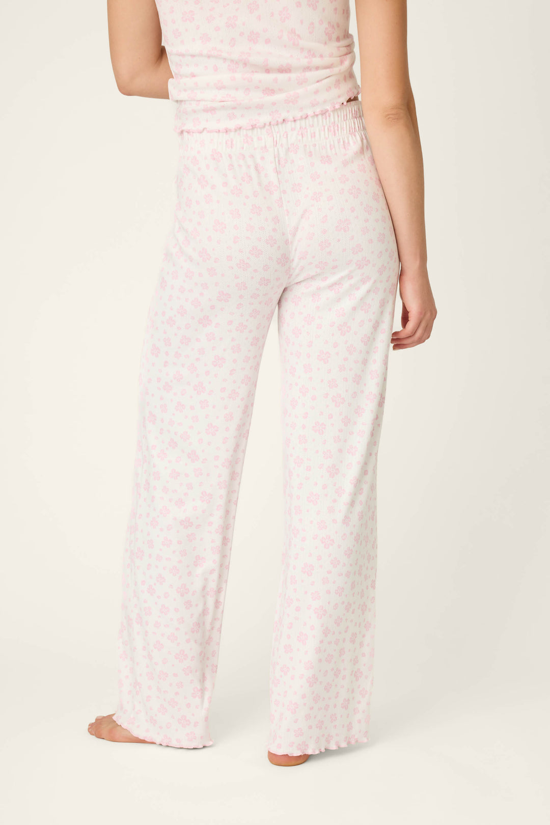 A person is barefoot, seen from the back, wearing P.J. Salvage Pretty Petals Sweet Pink Floral Print Open Leg Pants and a matching sleeveless floral top. The background is plain and light-colored.