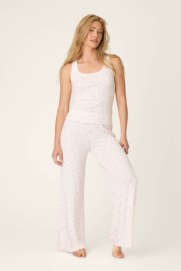 A woman with long blonde hair models the P.J. Salvage Pretty Petals Sweet Pink Floral Print Long PJ Set, featuring a light pink floral tank top and matching wide-leg pajama pants, as she stands barefoot against a plain light background.
