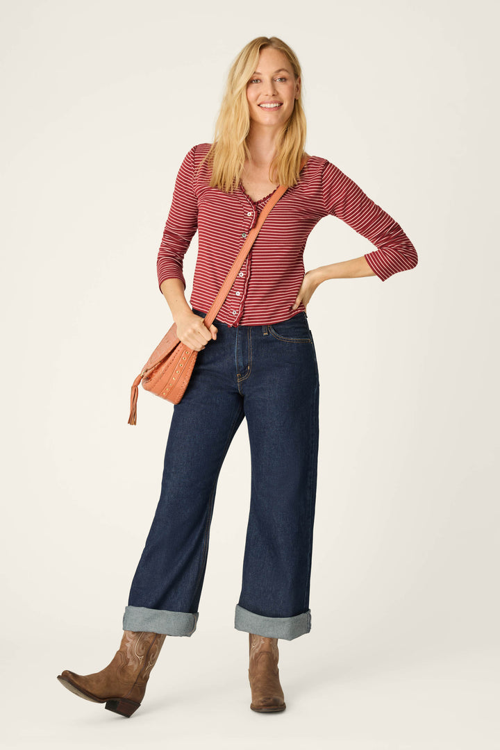 A smiling woman with blonde hair stands against a plain, light background wearing the P.J. Salvage Pretty Stripes Dark Wine Pointelle Cardigan. She pairs it with wide-cuffed blue jeans, brown boots, and carries a tan shoulder bag, standing confidently with one hand on her hip.