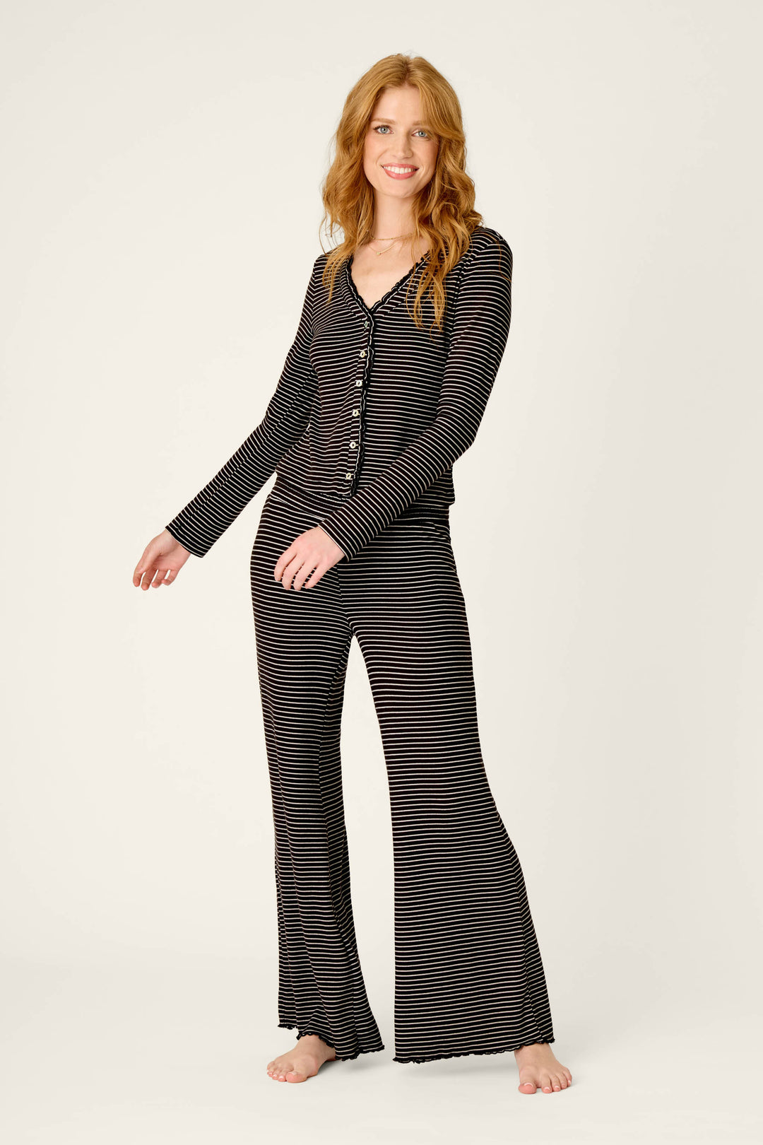 A person with long red hair smiles while wearing the Pretty Stripes Onyx Pointelle Cardigan by P.J. Salvage, standing barefoot against a plain light background.