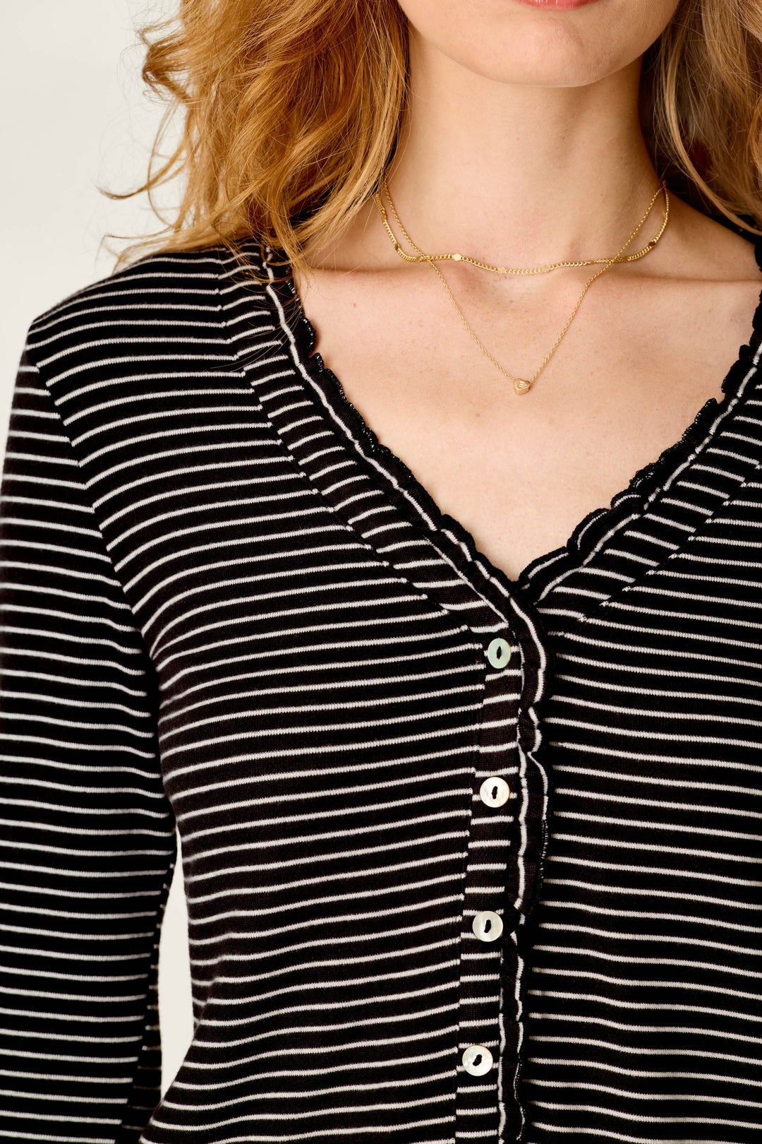 A woman with wavy blonde hair and two delicate gold necklaces is wearing the Pretty Stripes Onyx Pointelle Cardigan by P.J. Salvage—a black top featuring white horizontal stripes, a lace-trimmed V-neck, and buttons down the front.