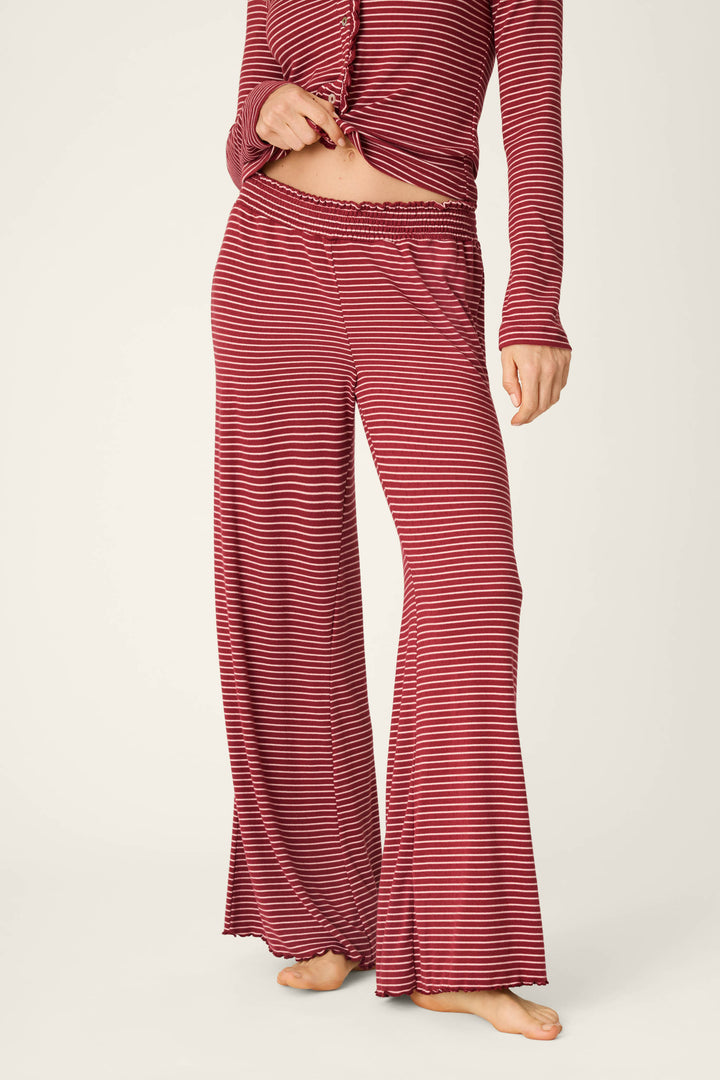 A person stands barefoot against a plain background, dressed in the P.J. Salvage Pretty Stripes Dark Wine Pointelle Pajama Pants with a matching long-sleeve top featuring red and black stripes; the top is partially unbuttoned and tied at the waist.