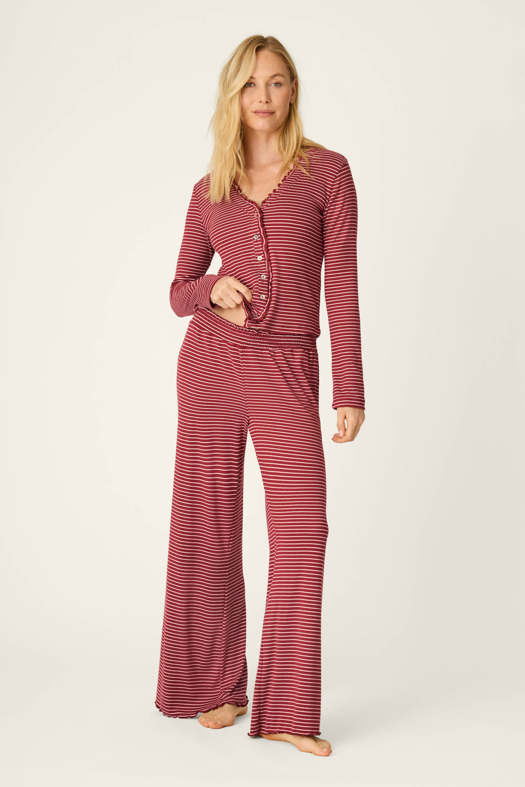 A woman poses barefoot against a plain, light background, wearing P.J. Salvage’s Pretty Stripes Dark Wine Pointelle Pajama Pants paired with a matching long-sleeve top featuring red and black stripes and wide-leg pants.