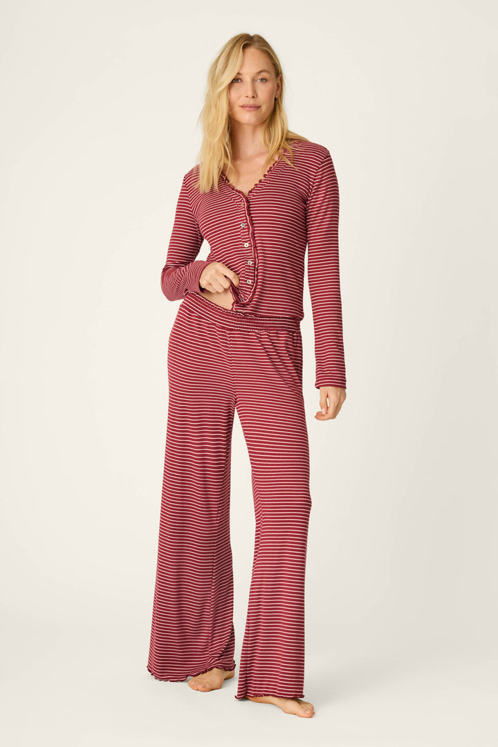 A woman poses barefoot against a plain, light background, wearing P.J. Salvage’s Pretty Stripes Dark Wine Pointelle Pajama Pants paired with a matching long-sleeve top featuring red and black stripes and wide-leg pants.