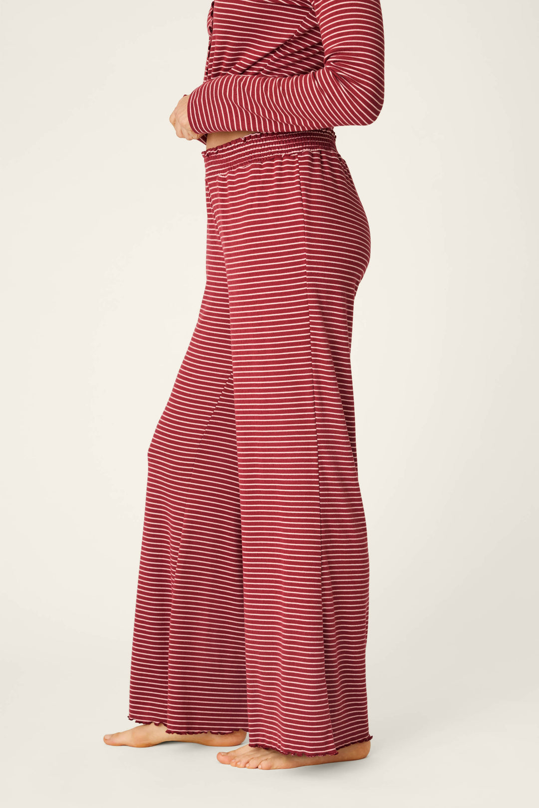 A person standing barefoot, shown from the side against a plain background, is wearing P.J. Salvage’s Pretty Stripes Dark Wine Pointelle Pajama Pants—a loose-fitting style featuring red and white horizontal stripes—paired with a matching long-sleeve top.