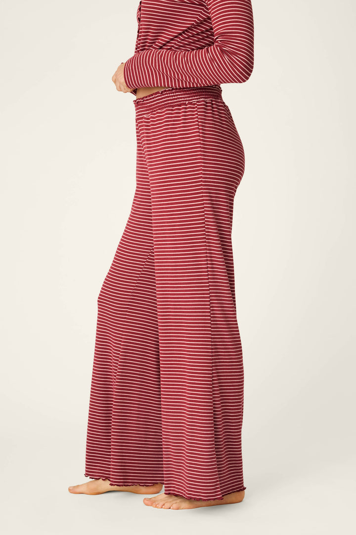 A person standing barefoot, shown from the side against a plain background, is wearing P.J. Salvage’s Pretty Stripes Dark Wine Pointelle Pajama Pants—a loose-fitting style featuring red and white horizontal stripes—paired with a matching long-sleeve top.