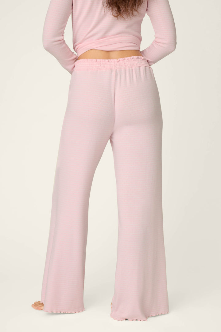 A person stands facing away, wearing P.J. Salvage’s Pretty Stripes Tender Pink Pointelle Pajama Pants. The light pink pants feature a subtle ribbed texture and ruffled hems, paired with a matching long-sleeve top. The background is plain and light-colored.