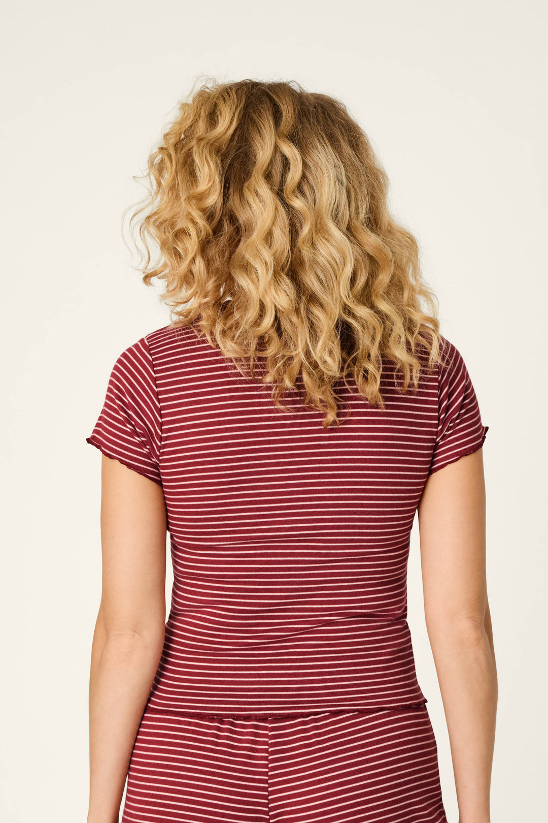 A person with curly blonde hair is seen from behind, wearing the Pretty Stripes Dark Wine Pointelle Short Sleeve Top by P.J. Salvage against a plain, light background.