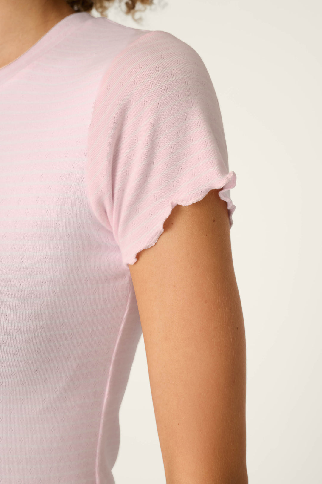 A person wearing the P.J. Salvage Pretty Stripes Tender Pink Pointelle Short Sleeve Top, featuring subtle pointelle detailing and a wavy lettuce-edge hem on the sleeve; only the shoulder, sleeve, and upper arm are visible.