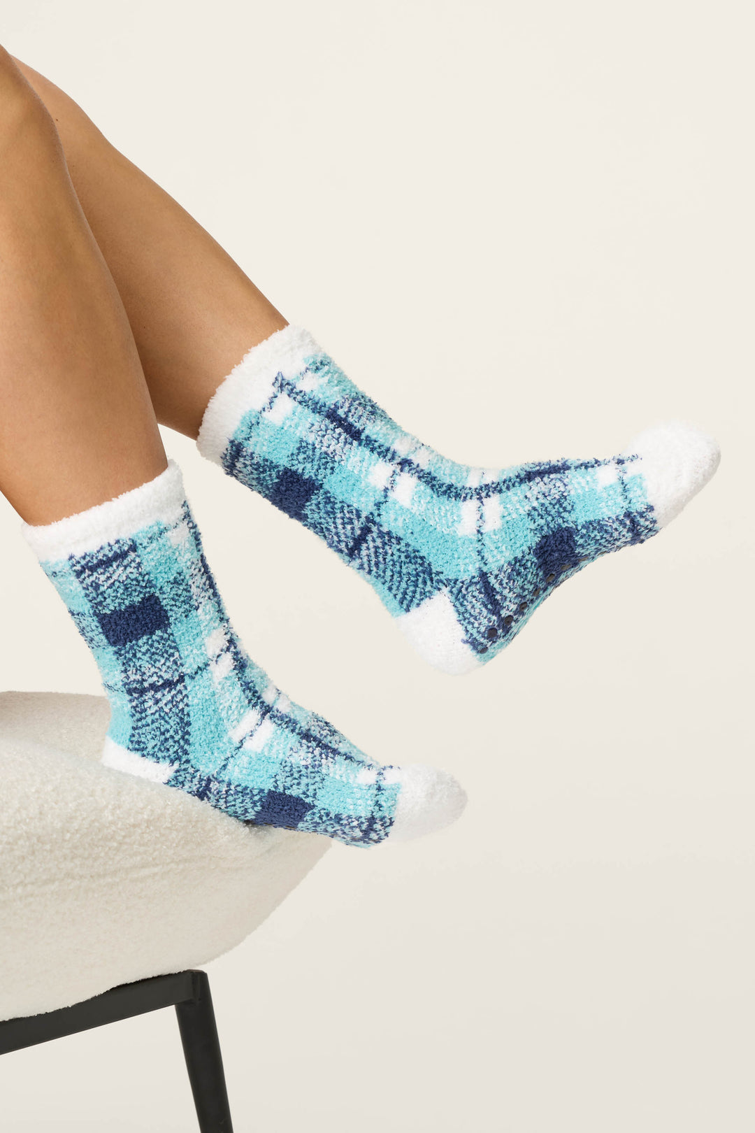 A person wearing P.J. Salvage Plaid About You Blue & Green Plaid Socks with white toes and cuffs sits on a white textured chair, legs stretched out against a plain light background.