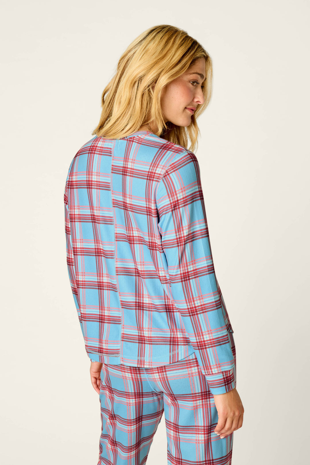 A woman with blonde hair wears the P.J. Salvage “Plaid About You” blue and red plaid print long sleeve top and matching pants, standing turned away and looking to the side against a plain light background.