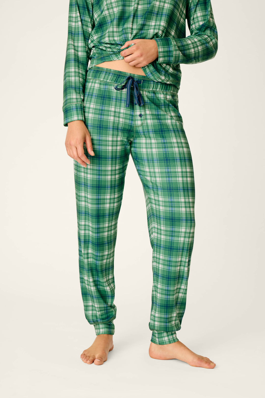 A person is shown barefoot against a plain light background, wearing P.J. Salvage’s Plaid About You Blue & Green Plaid Jogger Pajama Pants, which feature a drawstring waist and relaxed fit. Only the lower body and torso are visible.