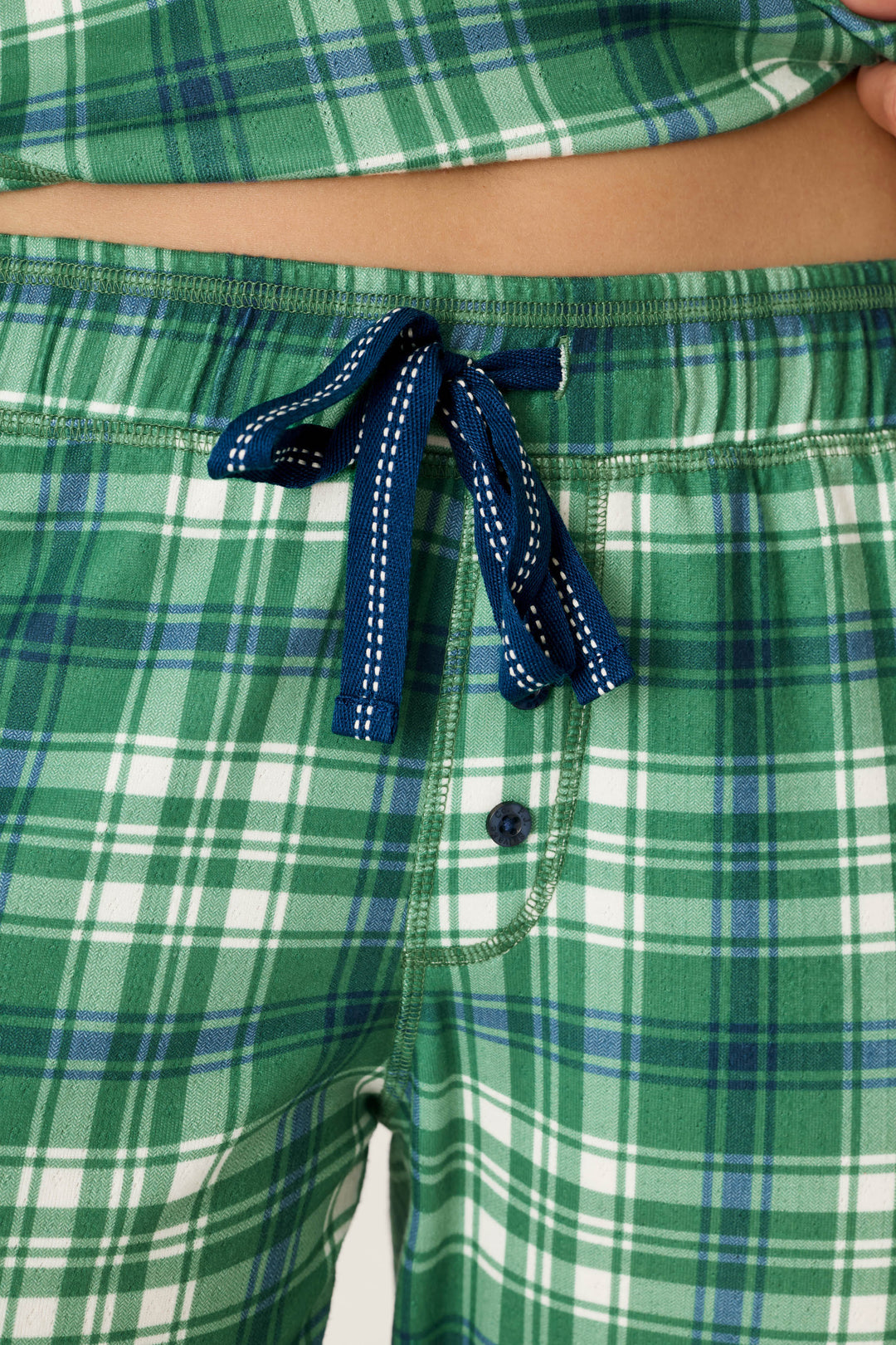 Close-up of a person wearing P.J. Salvage Plaid About You Blue & Green Plaid Jogger Pajama Pants, featuring a navy blue drawstring and button fly, with part of their bare midriff visible.