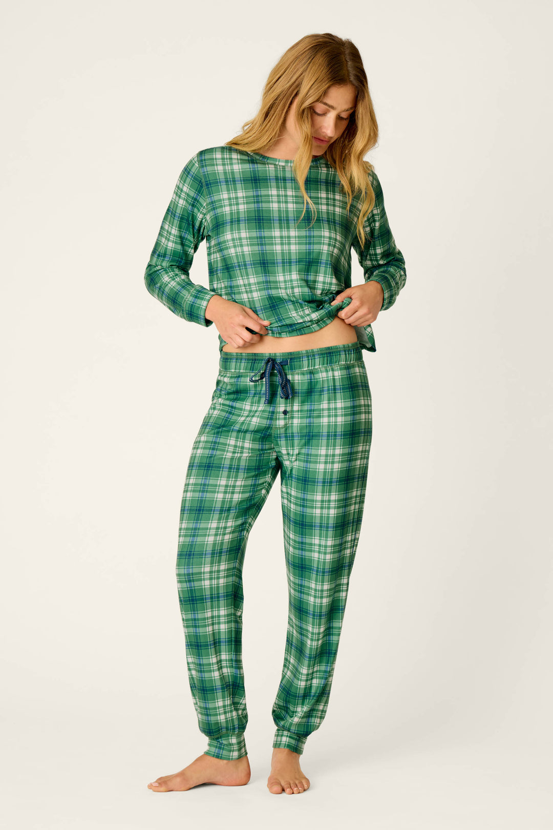 A woman stands barefoot against a plain light background, looking down as she adjusts her shirt while wearing the P.J. Salvage Plaid About You Blue & Green Plaid Jogger Pajama Pants.