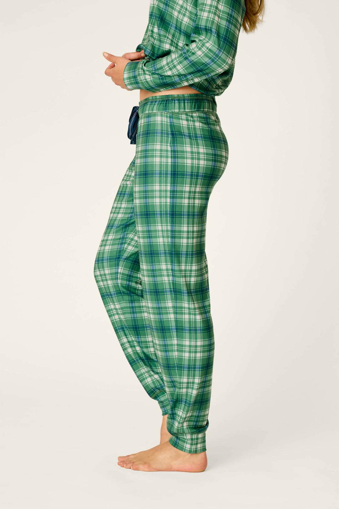 A person wearing P.J. Salvage Plaid About You Blue & Green Plaid Jogger Pajama Pants stands barefoot, photographed from the side against a plain light background. The image highlights the pajama pants.