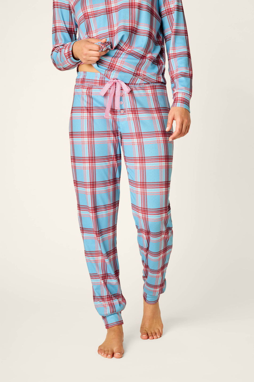A person stands barefoot on a light background, wearing P.J. Salvage’s Plaid About You Blue & Red Plaid Print Jogger Pajama Pants with a pink drawstring; the upper part of their face is out of frame.