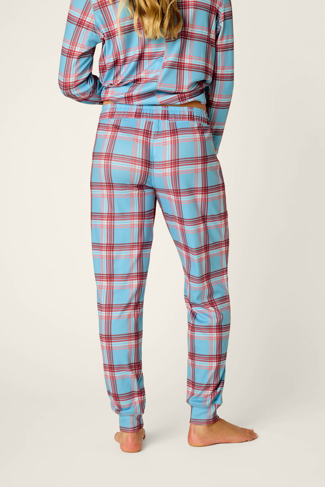 A person stands barefoot facing away, wearing the P.J. Salvage Plaid About You Blue & Red Plaid Print Jogger Pajama Pants. Their matching pajama top is partially visible, and their long hair cascades down their back.
