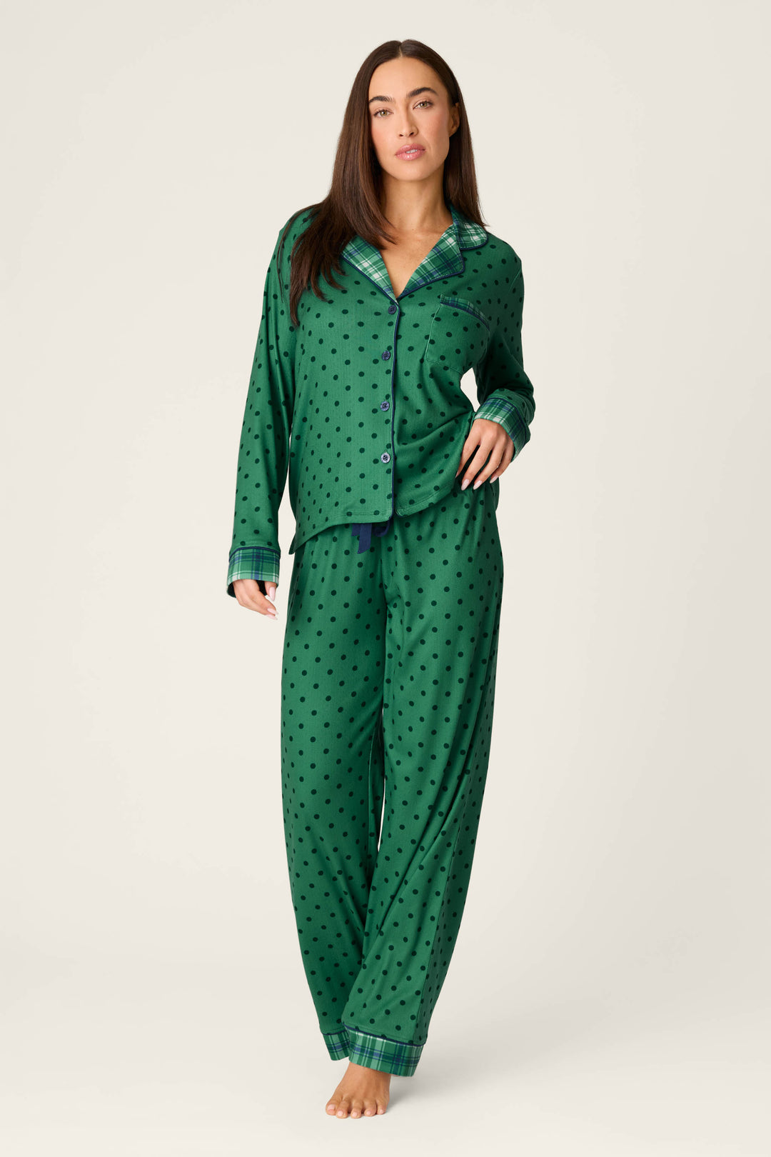 A woman stands against a plain light background wearing the Plaid About You Polka Dots & Plaid PJ Set by P.J. Salvage—green, long-sleeve pajamas with black polka dots and plaid trim—looking relaxed with one hand in her pocket.