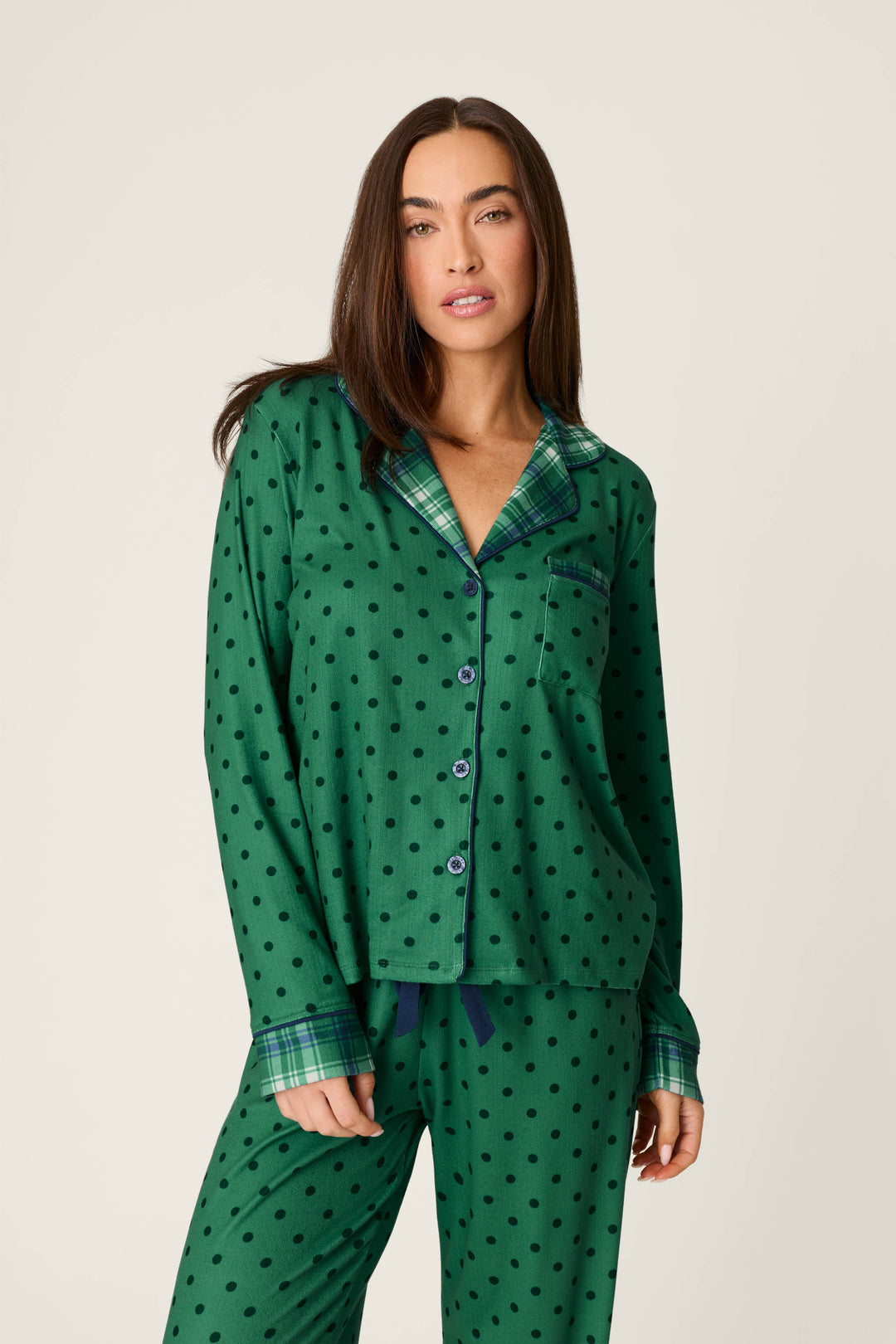 A woman wears the Plaid About You Polka Dots & Plaid PJ Set by P.J. Salvage, featuring green polka dot pajamas with plaid collar and pocket trim, standing against a plain light background and looking at the camera with a relaxed expression.