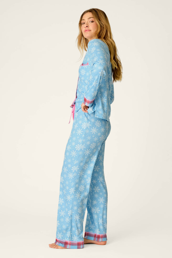 A woman models the P.J. Salvage Plaid About You Snowflakes & Plaid PJ Set, featuring light blue pajamas with white snowflakes and red plaid trim. She stands barefoot on a white background, turned sideways with one hand in her pocket, looking at the camera.