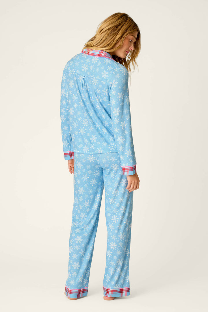A woman stands facing away, her blonde hair loose, wearing the P.J. Salvage Plaid About You Snowflakes & Plaid PJ Set—a light blue pajama set patterned with white snowflakes and finished with red plaid trim on the collar, cuffs, and pant hems—against a plain, light background.