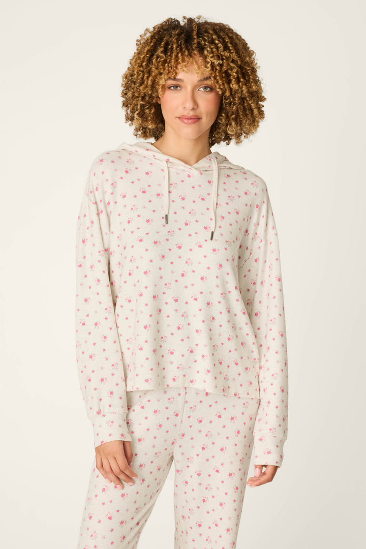 A woman wearing the P.J. Salvage Romantic Rose Ditsy Rose Print Hoodie with matching pants, featuring a delicate pink floral pattern, stands against a plain background and smiles softly.