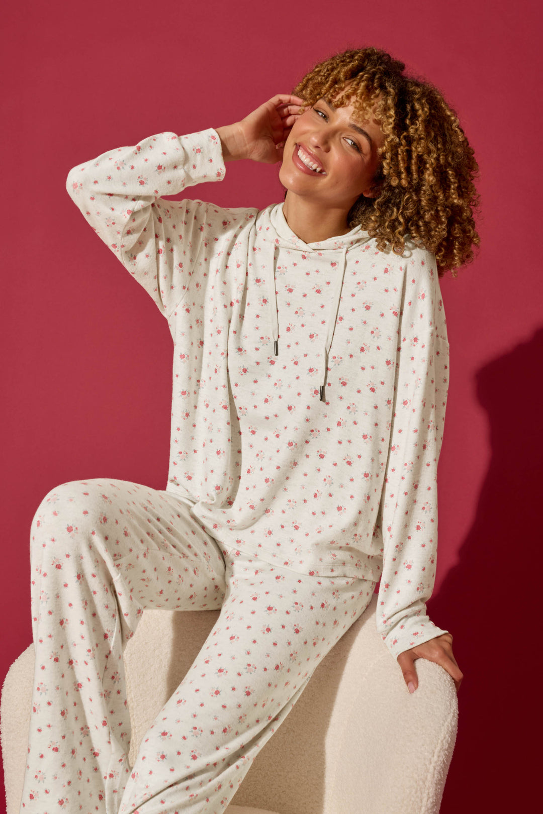 A smiling person with curly hair sits on a chair against a solid maroon background, wearing the P.J. Salvage Romantic Rose Ditsy Rose Print Hoodie and matching pants.