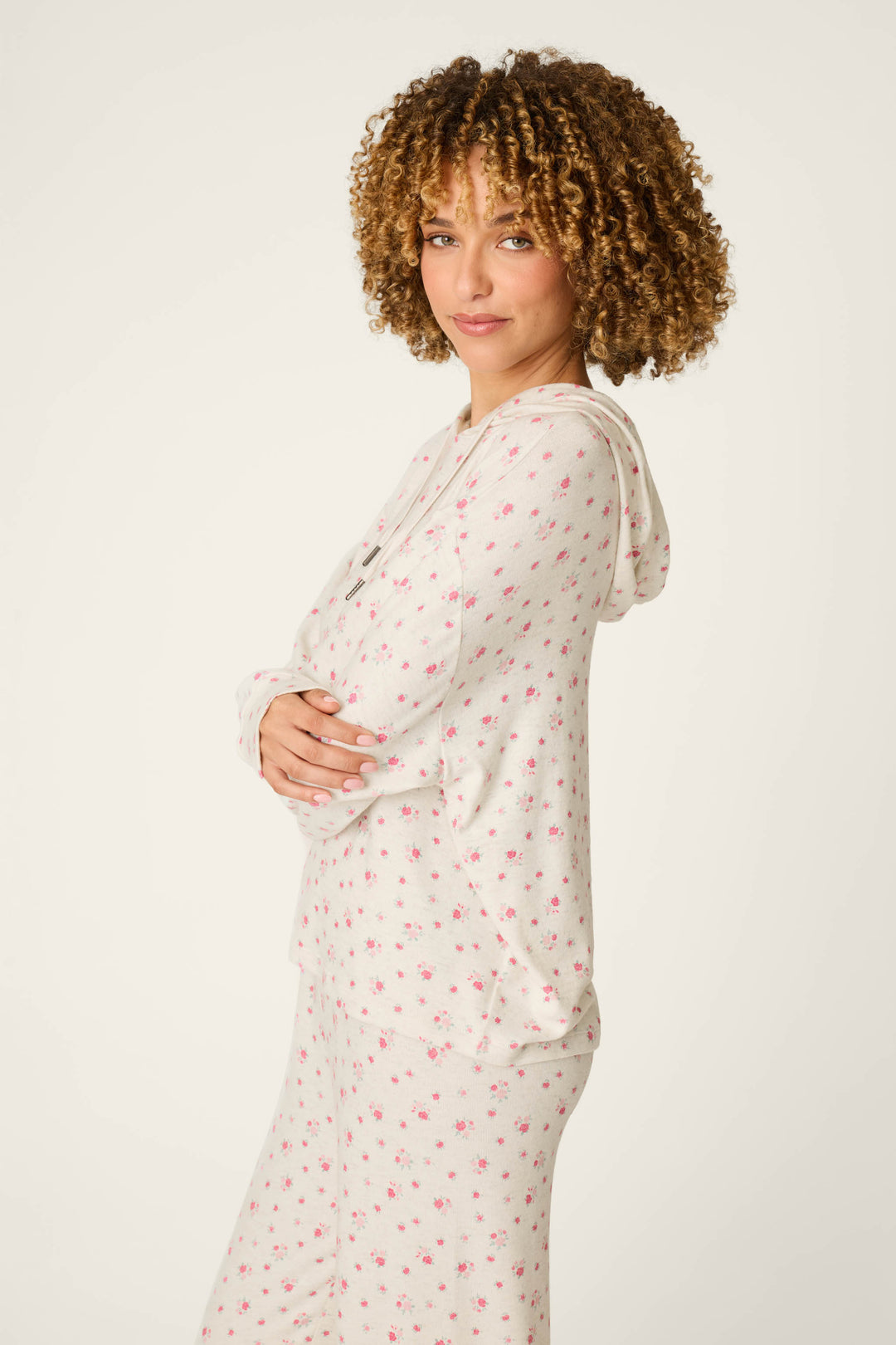 A woman with curly hair stands sideways with arms crossed and smiles gently against a plain light background, wearing the P.J. Salvage Romantic Rose Ditsy Rose Print Hoodie.