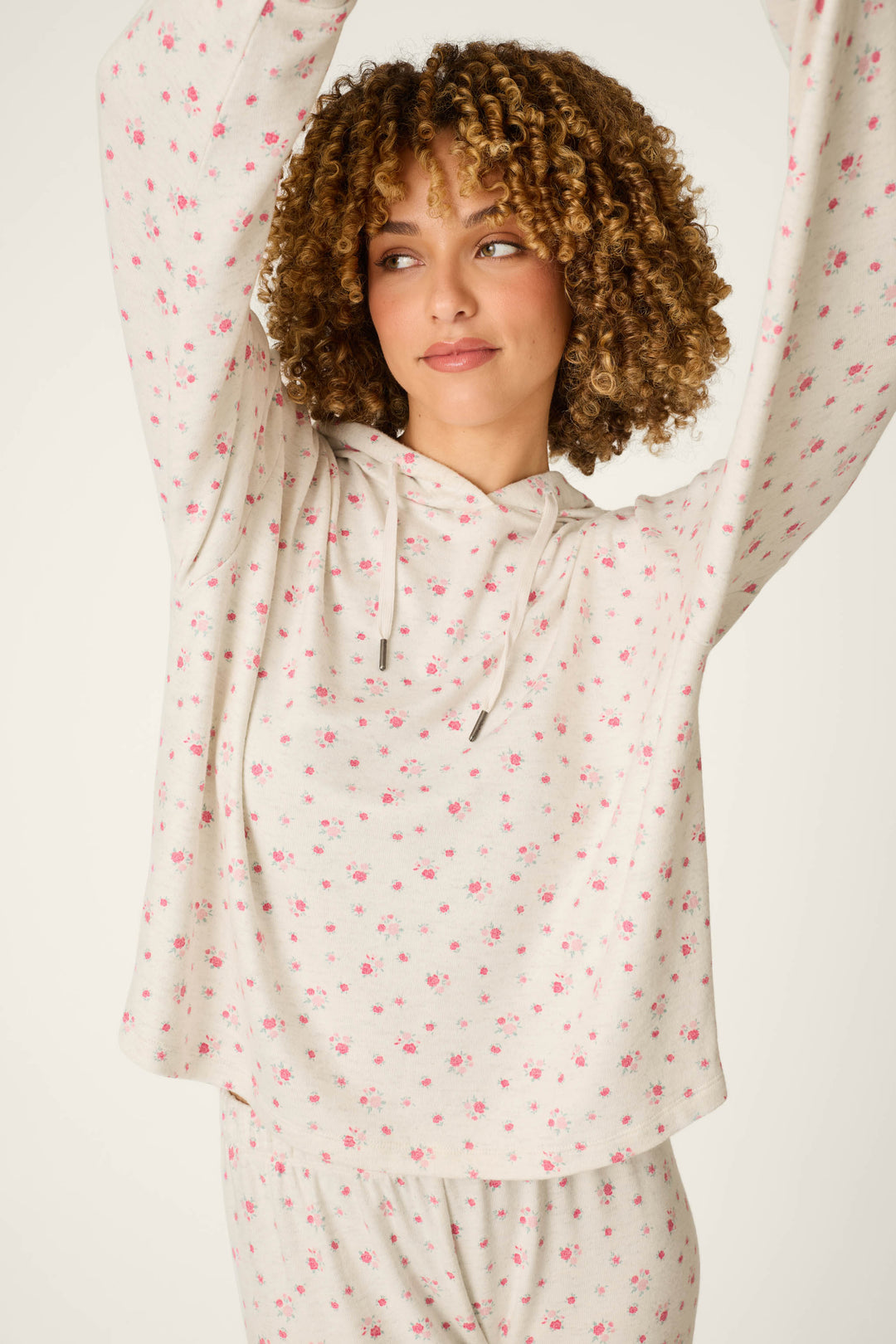 A person with curly hair wears the P.J. Salvage Romantic Rose Ditsy Rose Print Hoodie, looking to the side with arms raised against a plain background.