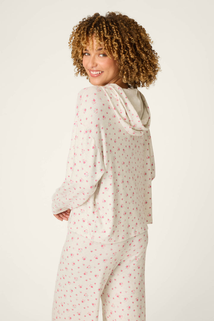 A woman with curly hair smiles while wearing the P.J. Salvage Romantic Rose Ditsy Rose Print Hoodie, a light-colored pajama set decorated with a pink floral design, facing away but looking over her shoulder.