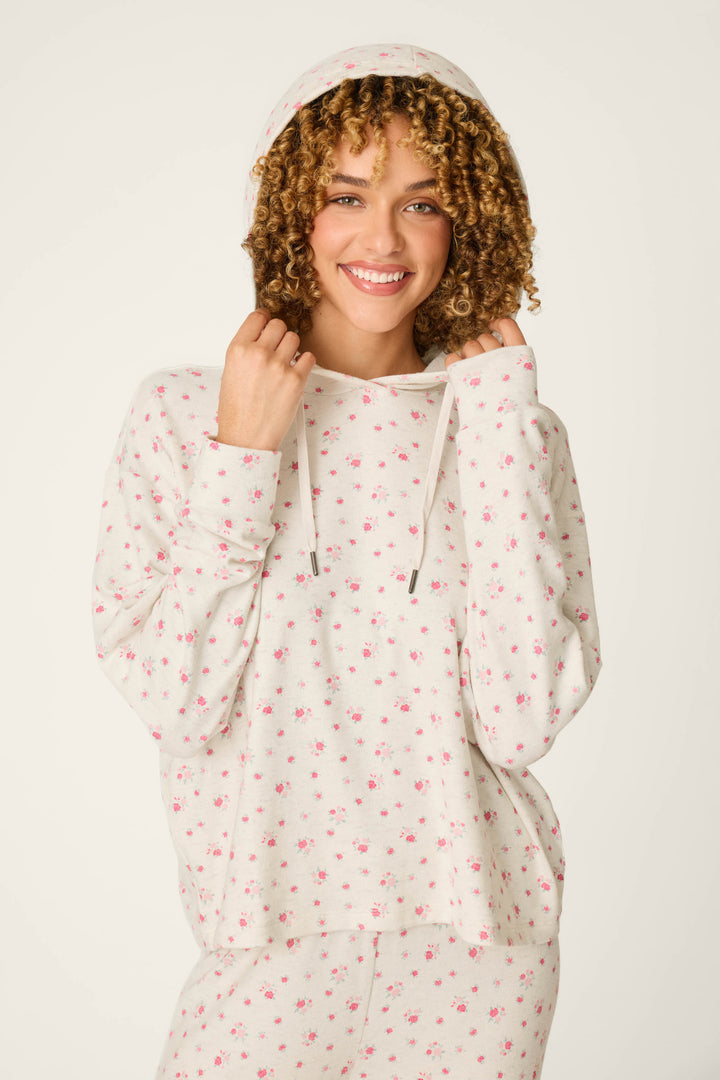 A woman smiles while wearing the P.J. Salvage Romantic Rose Ditsy Rose Print Hoodie, featuring delicate pink floral patterns, and holds the hood with both hands; matching pants are partially visible.