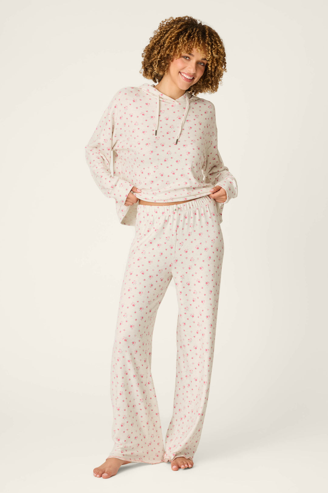 A smiling person with curly hair models the P.J. Salvage Romantic Rose Ditsy Rose Print Pajama Pants, featuring a cozy cream-colored design with a delicate rose print. They pair the loose pajama pants with a matching long-sleeve top and stand barefoot against a plain light background.